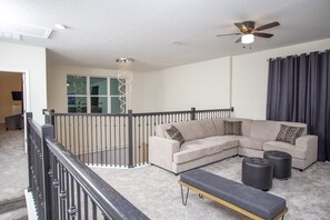 A living room with a beige sectional sofa, two ottomans, a bench, a ceiling fan, and a staircase with a railing. - Fabulous 5 Bedrooms Close to Disney at Champions Gate Resort 8988 (Kissimmee)