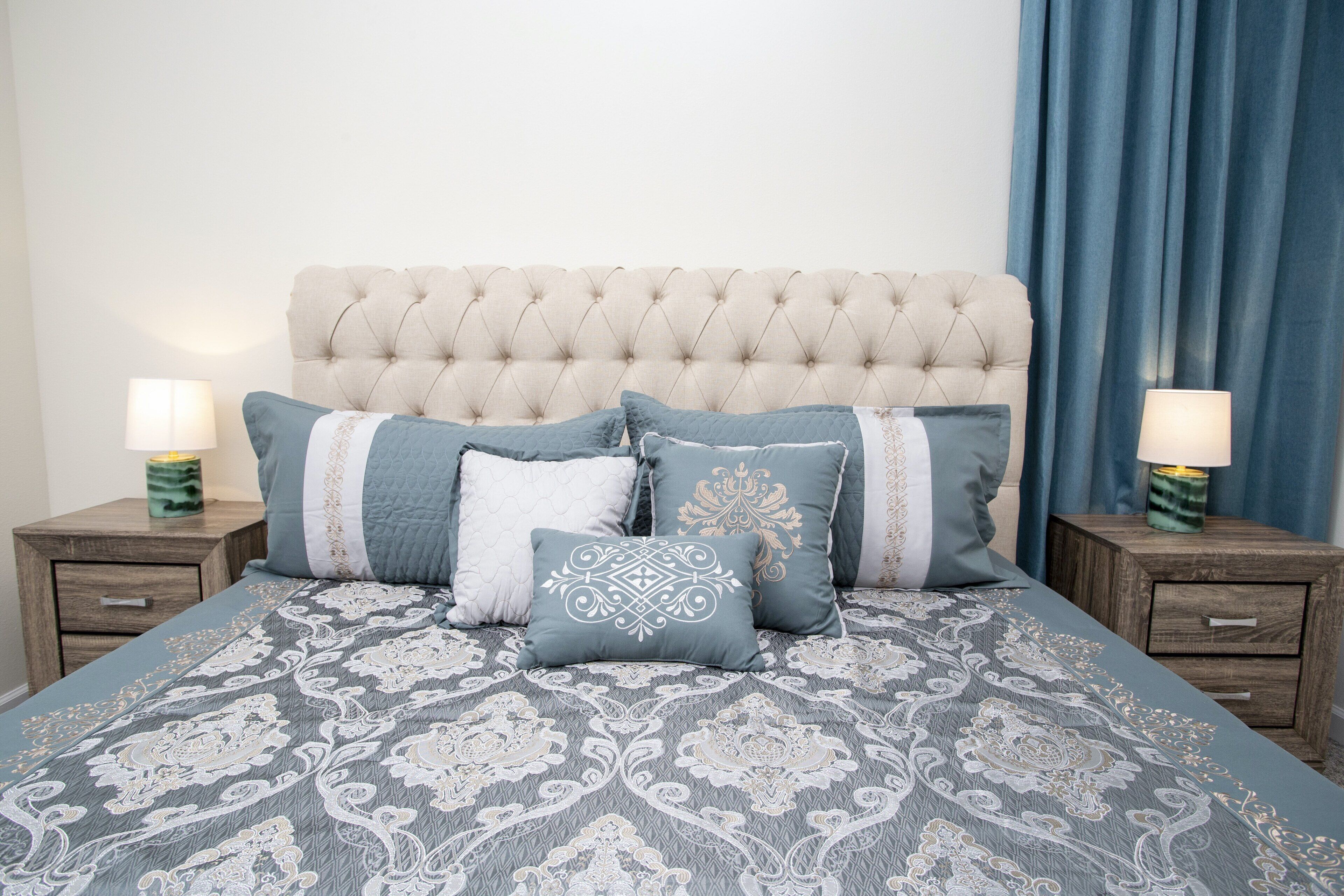 Una cama con cabecera acolchada, varios almohadones y una colcha estampada. Hay dos mesitas de noche con lámparas y un cajón pequeño.