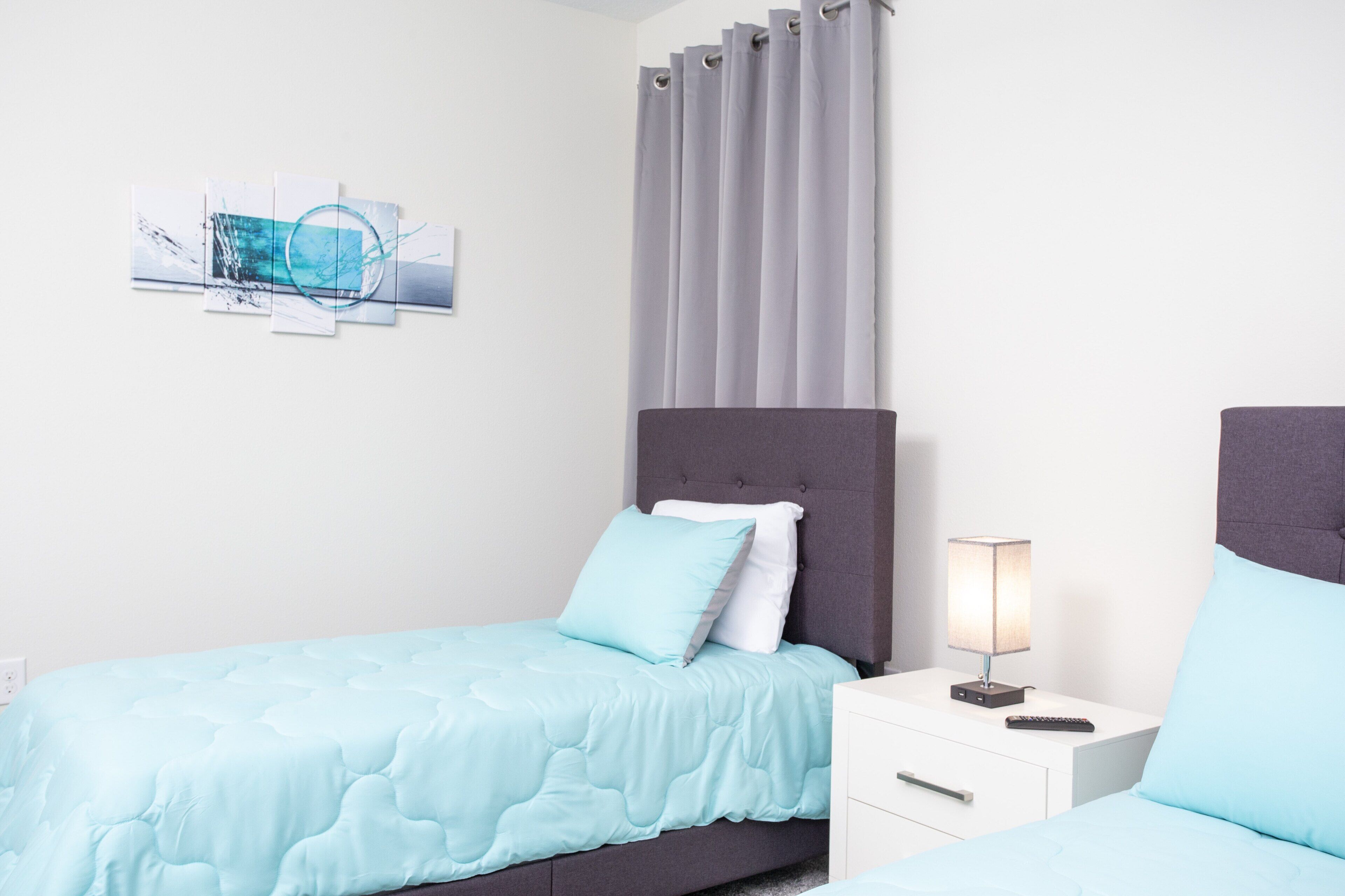 Un dormitorio moderno con cabecera gris, colcha azul claro, mesita de noche blanca con lámpara y una obra de arte en la pared.