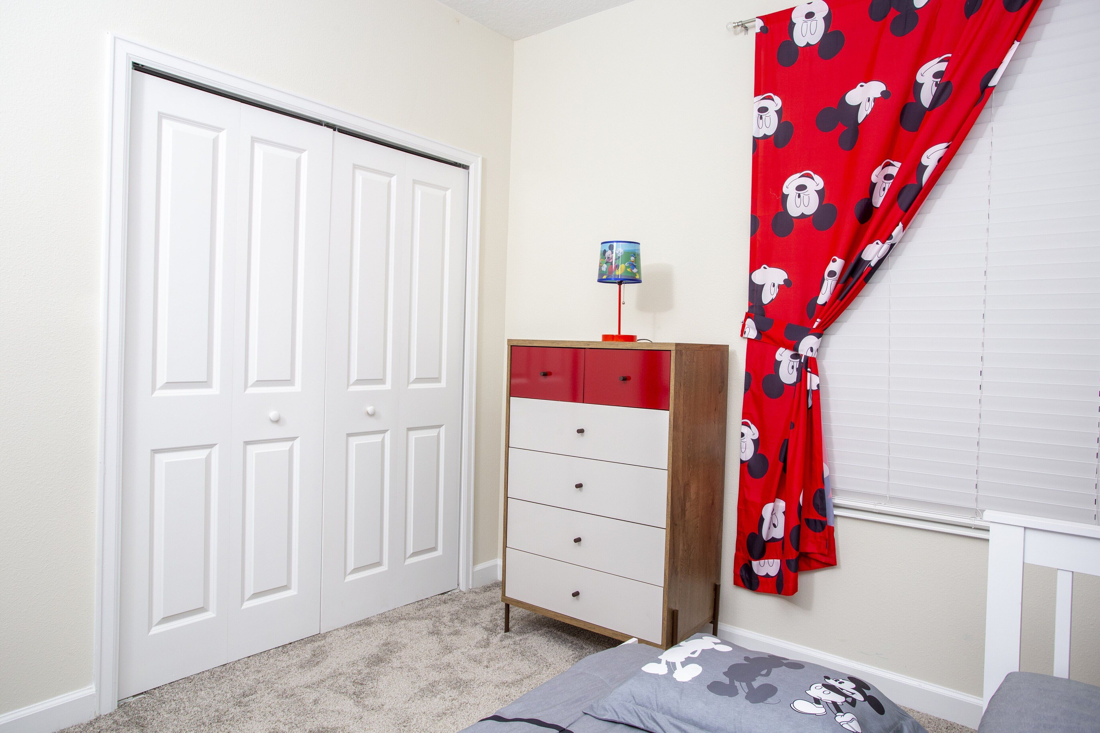 Ein Schlafzimmer mit einer weißen Tür, einem roten und weißen Schminktisch und einem Vorhang mit Mickey Mouse Motiv. Das Bett ist mit einem Mickey Mouse Kissen ausgestattet.