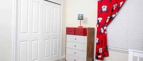 Ein Schlafzimmer mit einer weißen Tür, einem roten und weißen Schminktisch und einem Vorhang mit Mickey Mouse Motiv. Das Bett ist mit einem Mickey Mouse Kissen ausgestattet.