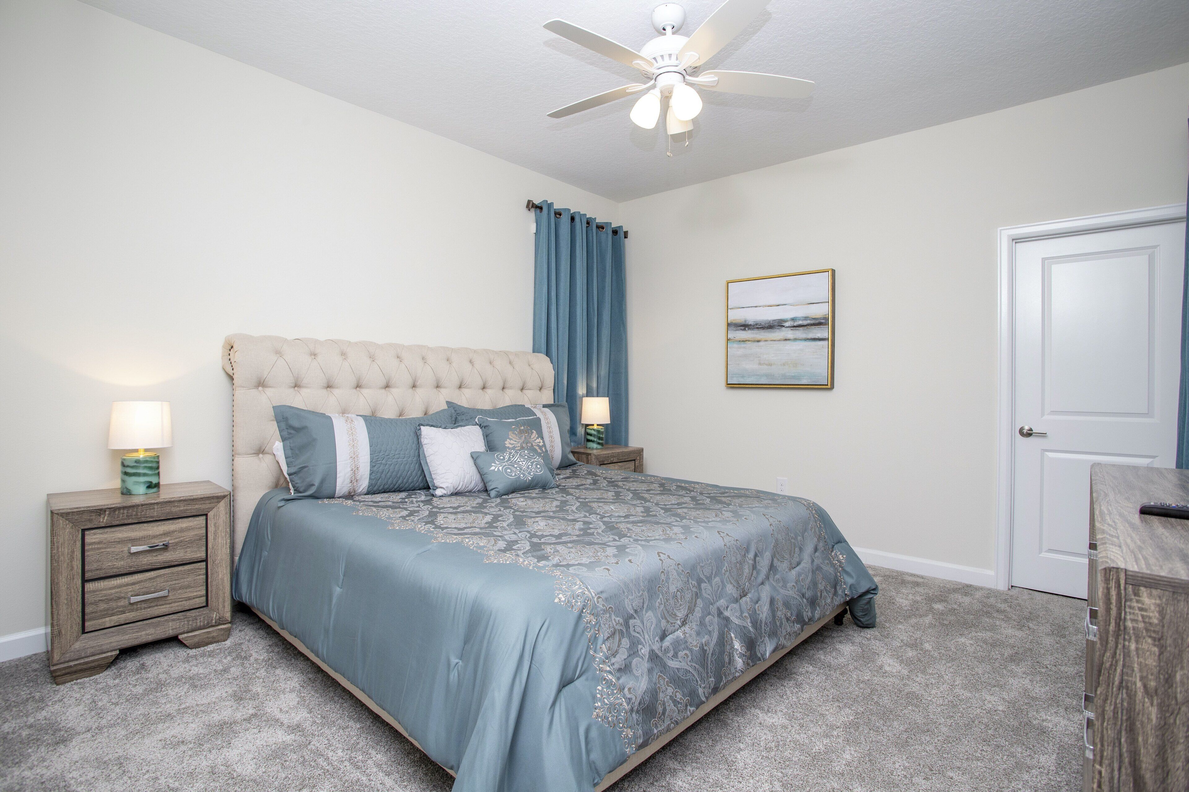 A bedroom with a tufted headboard, a bed with a blue patterned bedspread, two bedside tables with lamps, a ceiling fan, a door, and a framed picture on the wall.