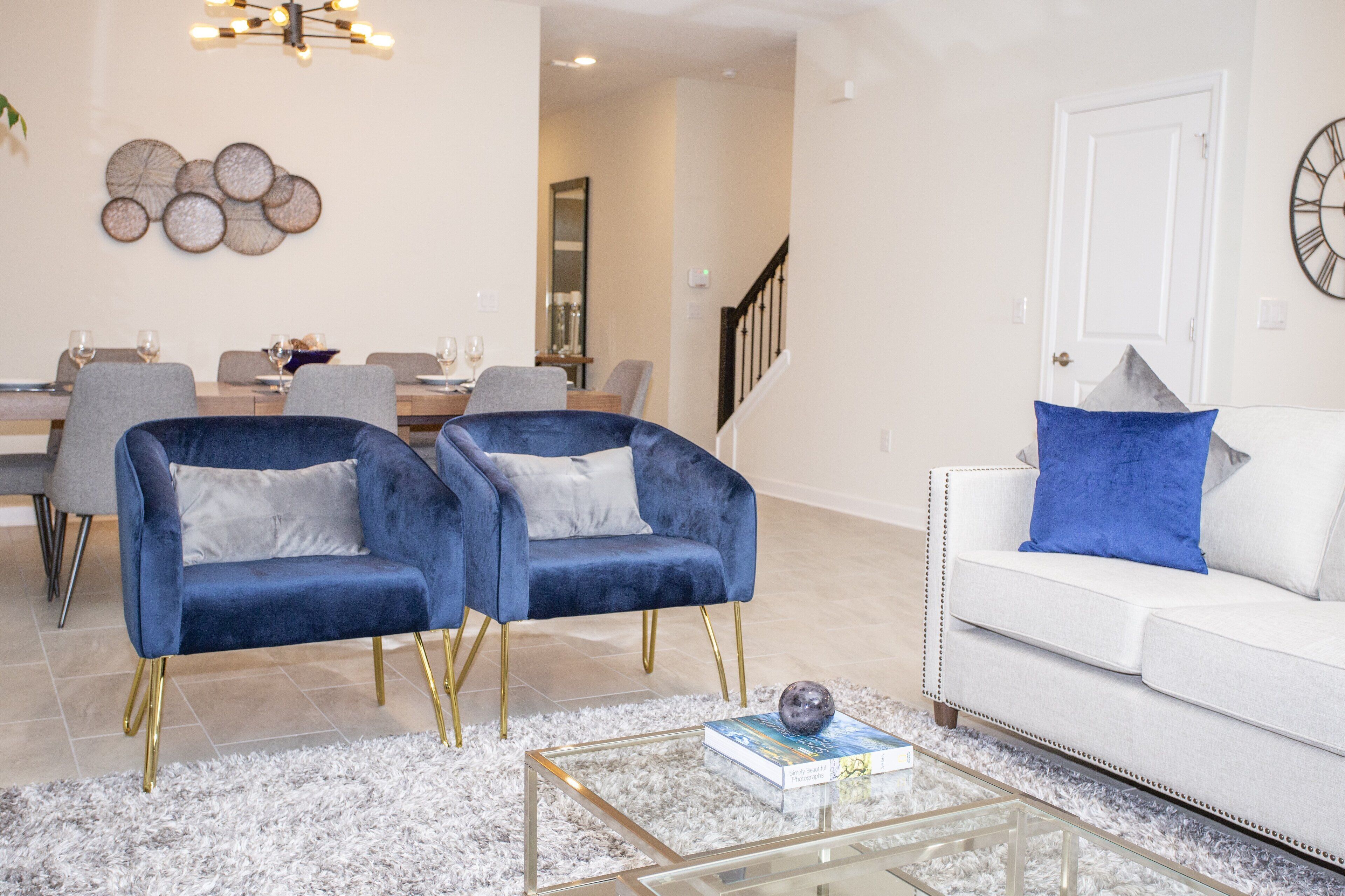A modern living room with a white sofa, two blue armchairs, a glass coffee table, and a dining area in the background.