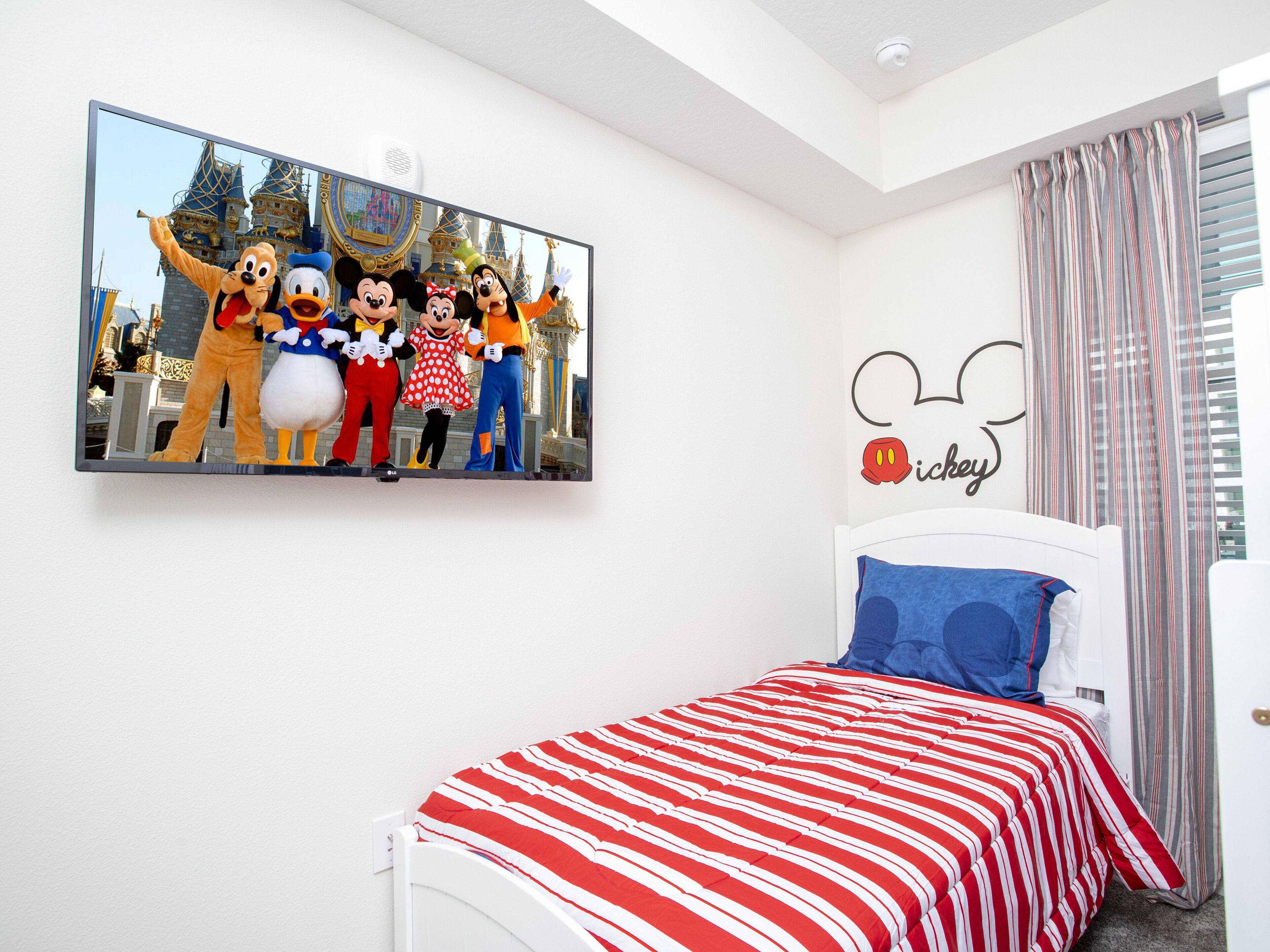 A hotel room with a bed featuring a red and white striped bedspread, a Mickey Mouse wall decal, and a large poster of Disney characters on the wall.