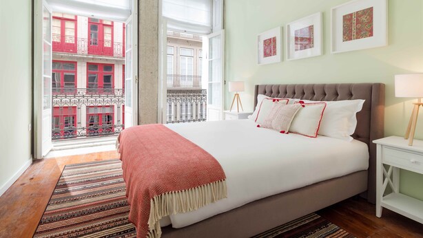 Een slaapkamer met een groot bed, uitzicht op een rode gevel door een raam, en een klein nachtkastje met een lamp.