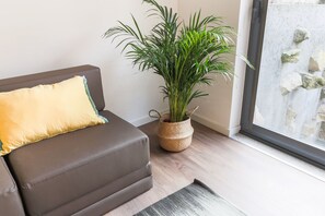 Un salon moderne avec un canapé en cuir marron, un coussin jaune, un pot de fleurs en osier et une vue sur un mur en pierre à travers une porte vitrée.