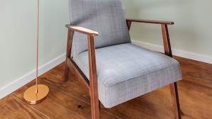 Una habitación con piso de madera, un sillón tapizado en gris, una lámpara de pie y un ventanal con persiana blanca abierta.