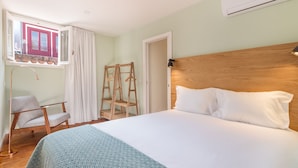 Un dormitorio con cama, una silla, un armario de madera, una ventana con cortinas y un equipo de aire acondicionado.