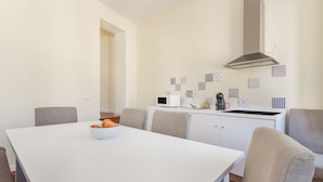 Una cocina moderna con armarios blancos, un microondas, un hervidor y una estufa a gas. Hay una mesa de comedor con sillas y un cuenco de fruta encima.