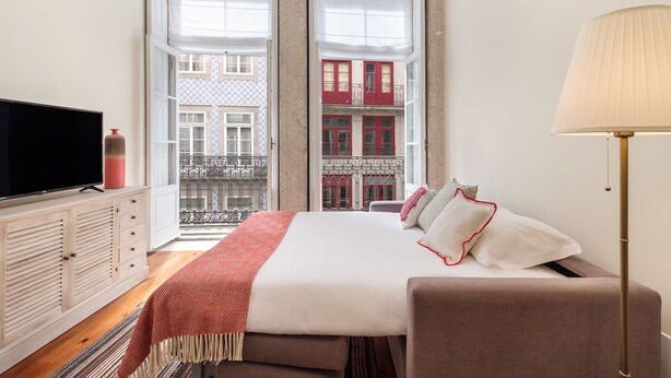 Een slaapkamer met een groot bed, een televisie op een kast, en uitzicht op een gebouw met balkons.