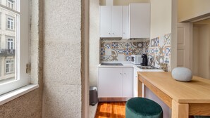 A compact kitchen with white cabinets, a tiled backsplash, a microwave, and a small wooden table with a green ottoman. - Nada02 · Faisama #2 Studio Next to Ribeira (Porto)