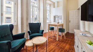 A room with a wooden floor, a green sofa, a chair, a small round table, a TV, and a cabinet with a bowl of apples. - Nada02 · Faisama #2 Studio Next to Ribeira (Porto)