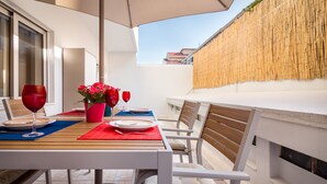 Un espace de restauration en plein air avec une table dressée pour deux personnes, deux verres à vin, un vase avec une fleur rose et un mur en bambou.