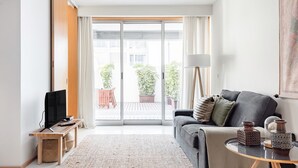 A modern living room with a sofa, a TV on a wooden stand, a coffee table with decorative items, and a view of a balcony with plants. - Mat001 · Bohemian Vibe at a Fresh Coastal Getaway (Matosinhos)