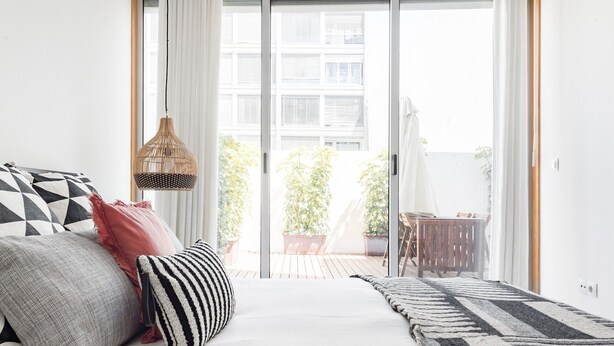 A bedroom with a bed, pillows, a hanging light fixture, and a view of a balcony with plants and a table. - Mat001 · Bohemian Vibe at a Fresh Coastal Getaway (Matosinhos)
