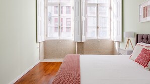 A bedroom with a bed, a bedside table, a lamp, and a window with curtains. - Nada01 · Cute and Simple Apartment in the Historic District (Porto)