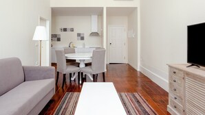 A compact living space with a sofa, dining table, chairs, a TV, and a kitchen area with a white door and patterned wall art. - Nada01 · Cute and Simple Apartment in the Historic District (Porto)