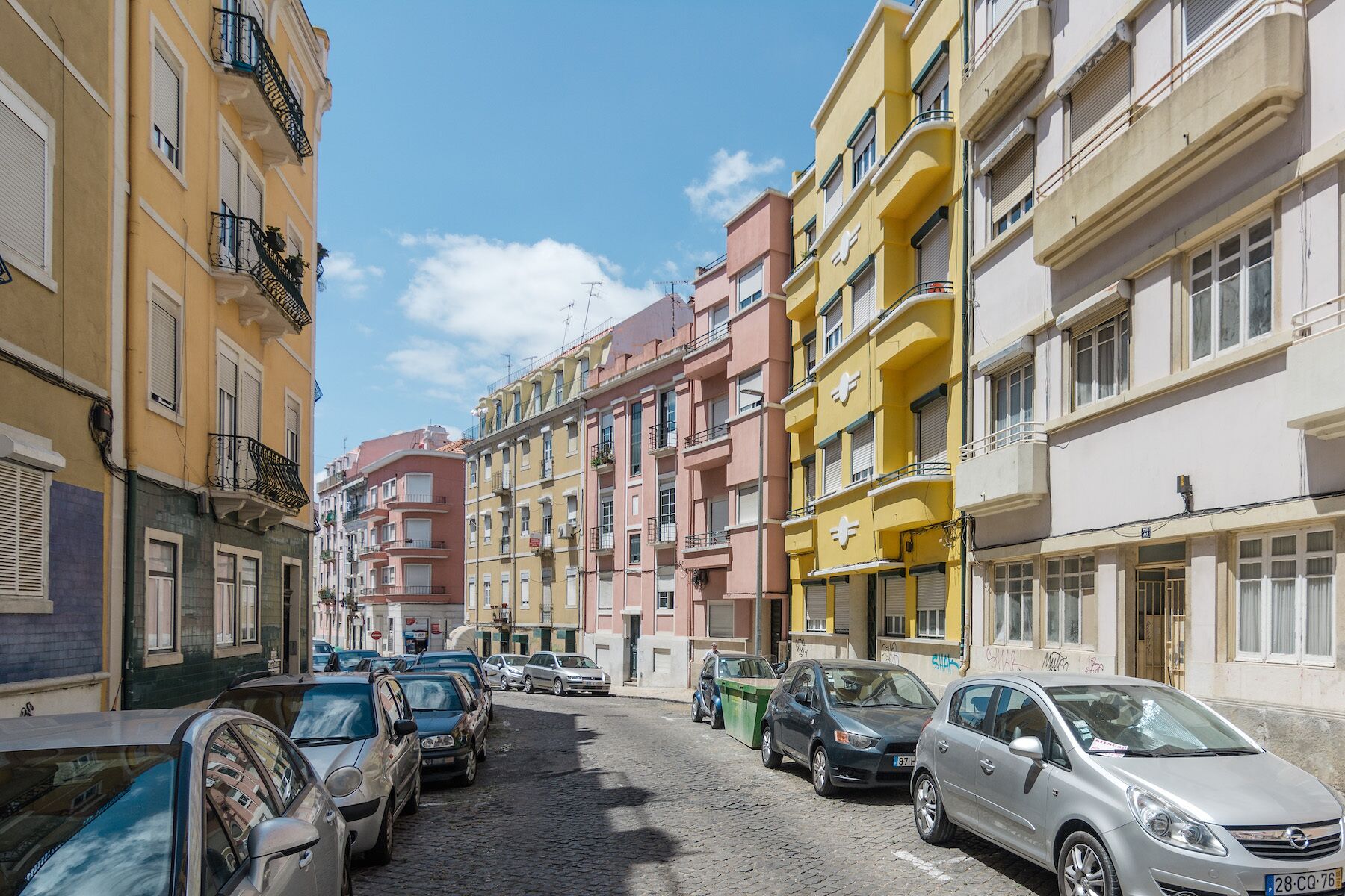 A narrow cobblestone street lined with multi-story buildings, parked cars, and balconies.