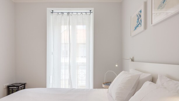 Ein schlicht eingerichtetes Schlafzimmer mit einem großen Bett, einem Fenster mit durchsichtigen Vorhängen und einem kleinen Nachttisch mit Lampe.