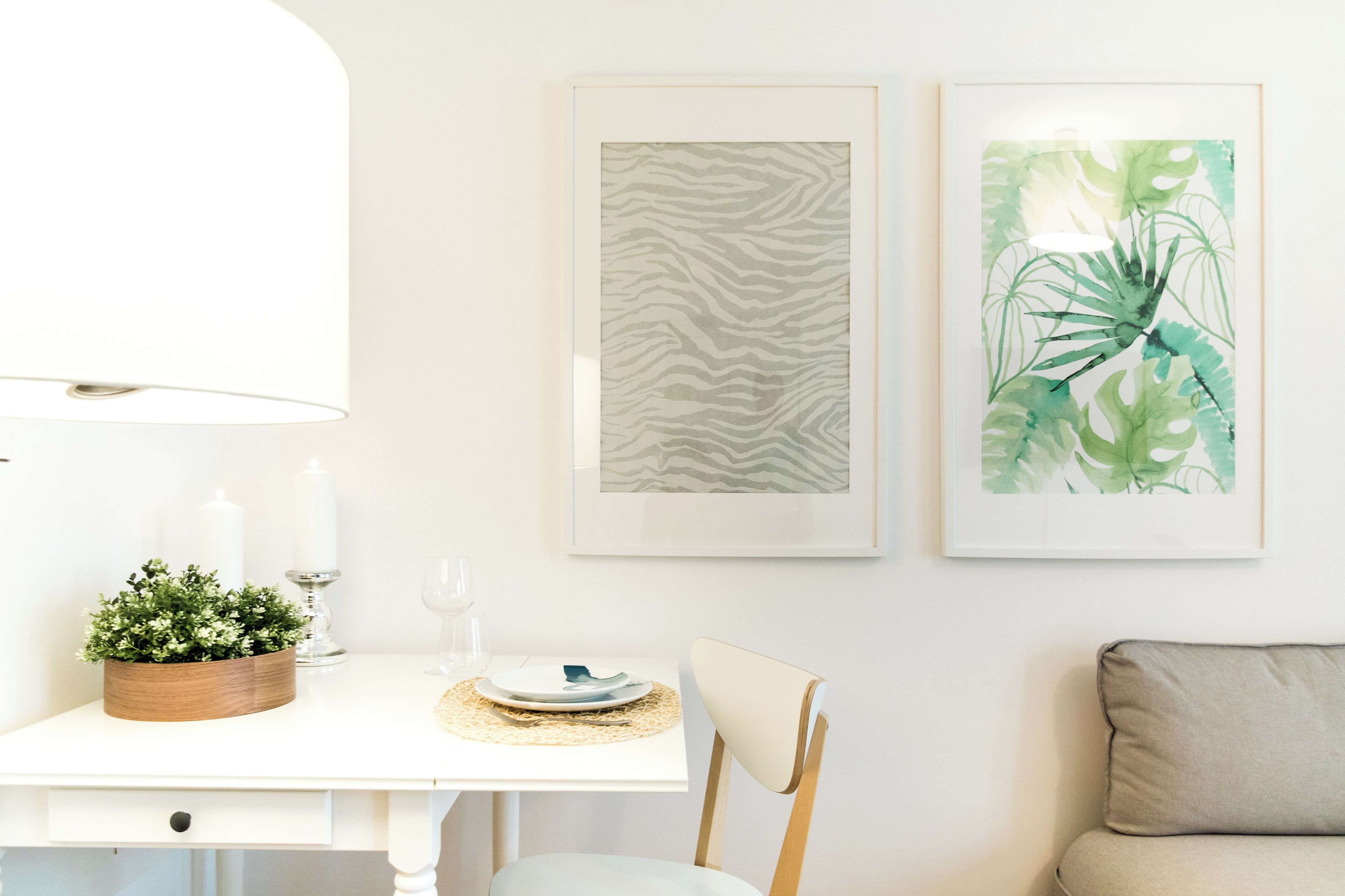 Een eetgedeelte met een witte tafel, een houten bijzettafeltje met kaarsen, en twee kunstwerken in lijsten aan de muur.
