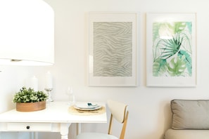 Un coin repas avec une table blanche, une petite table d’appoint en bois ornée de bougies et deux œuvres d’art encadrées accrochées au mur.