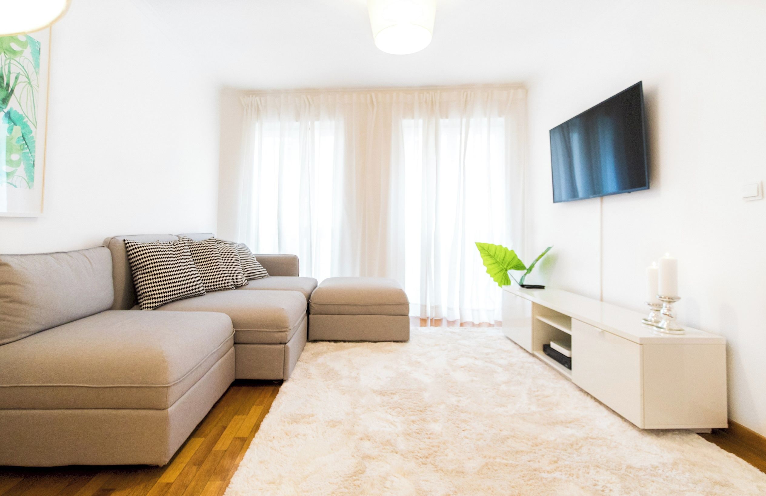 Una sala de estar moderna con un sofá color beige, un televisor de pantalla plana y un amplio ventanal con cortinas translúcidas.