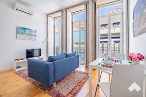 A modern living room with a blue sofa, a TV on a white stand, a dining table with chairs, and a balcony with a view of buildings. - Mrtir1 · Bright Apartment Downtown Porto (Porto)