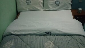 A bed with a grey quilt and pillows with the 'Addis' logo, a wooden headboard, and a small bedside table with a can. - Addis Lodge (Gilgil)