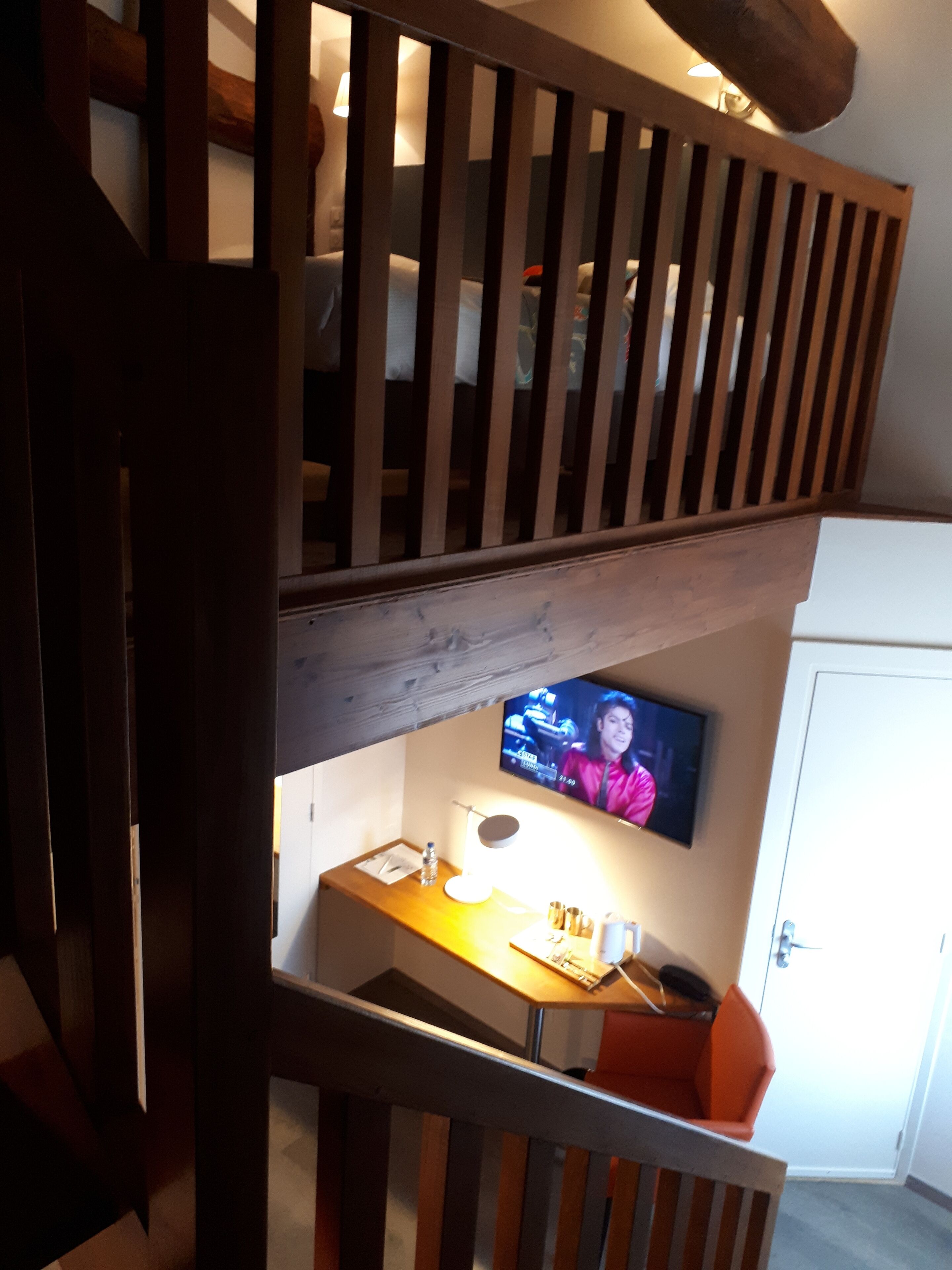 A staircase with wooden railings, a television mounted on the wall, a desk with a lamp, and an orange chair.