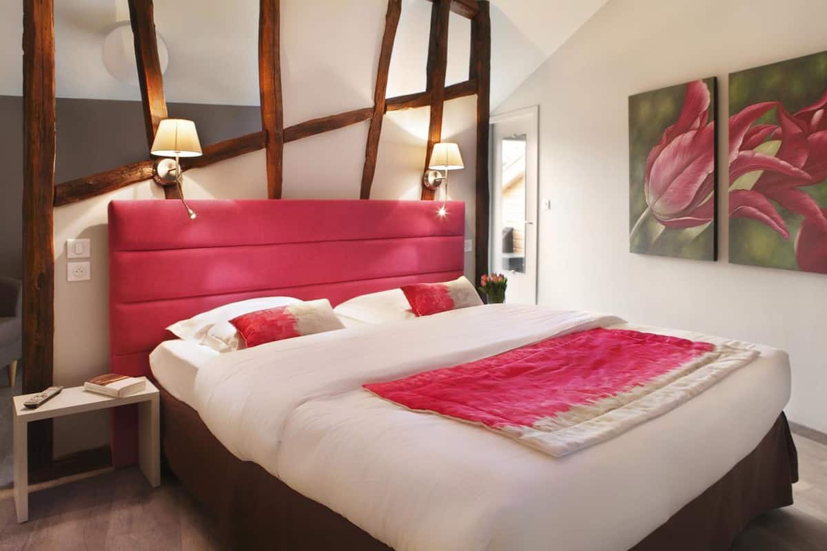 Een bed met een rode hoofdbord, witte beddengoed en een rode plaid. Er staan twee bedlampen en een nachtkastje met een boek op.
