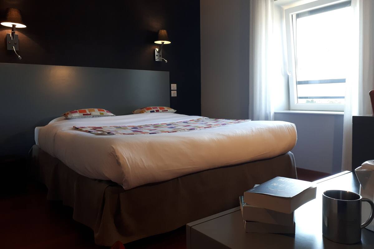 Een hotelkamer met een bed, nachtlampjes, een raam met gordijnen, een bureau met een mok en een boek.