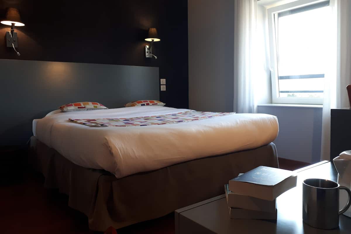 A hotel room with a bed, bedside lamps, a window with curtains, a desk with a mug, and a book.