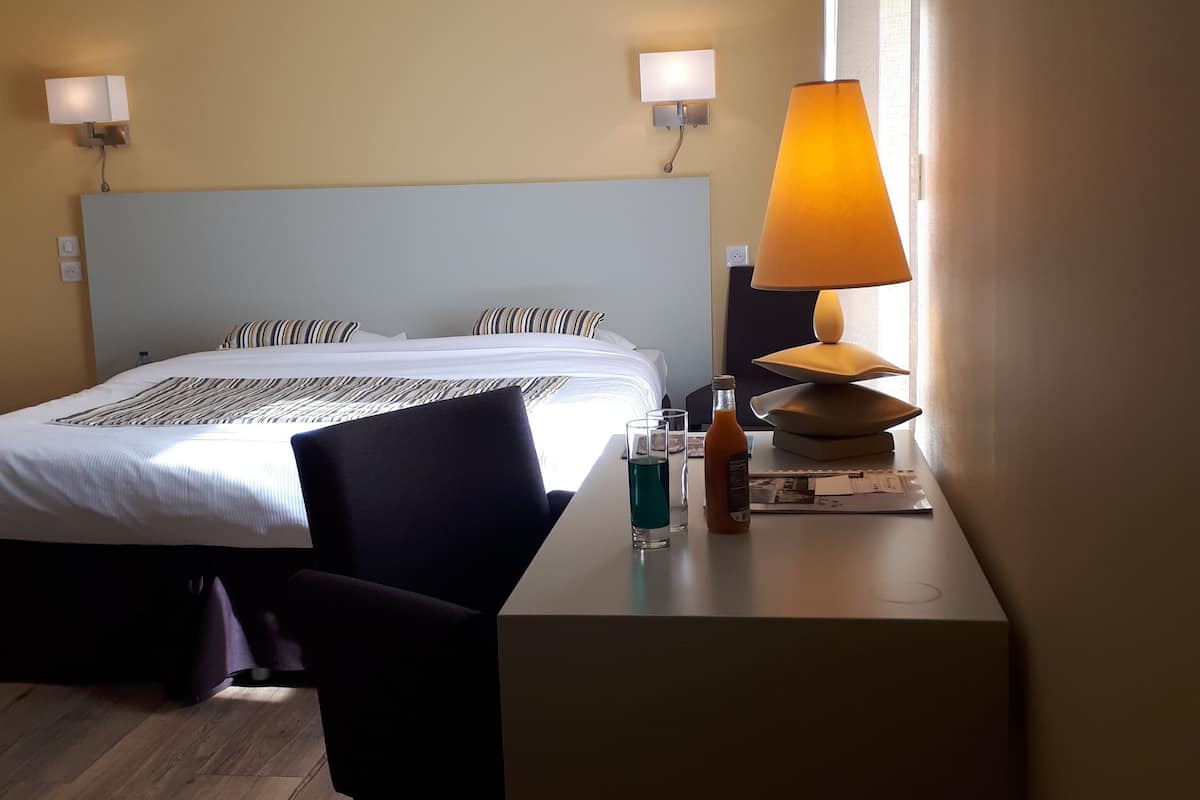 Een hotelkamer met een bed, nachtlampjes, een bureau met lamp, een stoel en een raam.