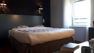 A hotel room with a bed, bedside tables, a window with curtains, and wall-mounted lamps.