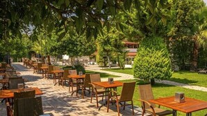 An outdoor seating area with wooden tables and chairs, surrounded by lush greenery and a pathway leading to a building. - Rose Resort Hotel (Kemer)