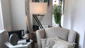Un salon chaleureux avec un fauteuil gris, une petite table d’appoint avec une lampe, un tableau encadré au mur et une plante en pot.