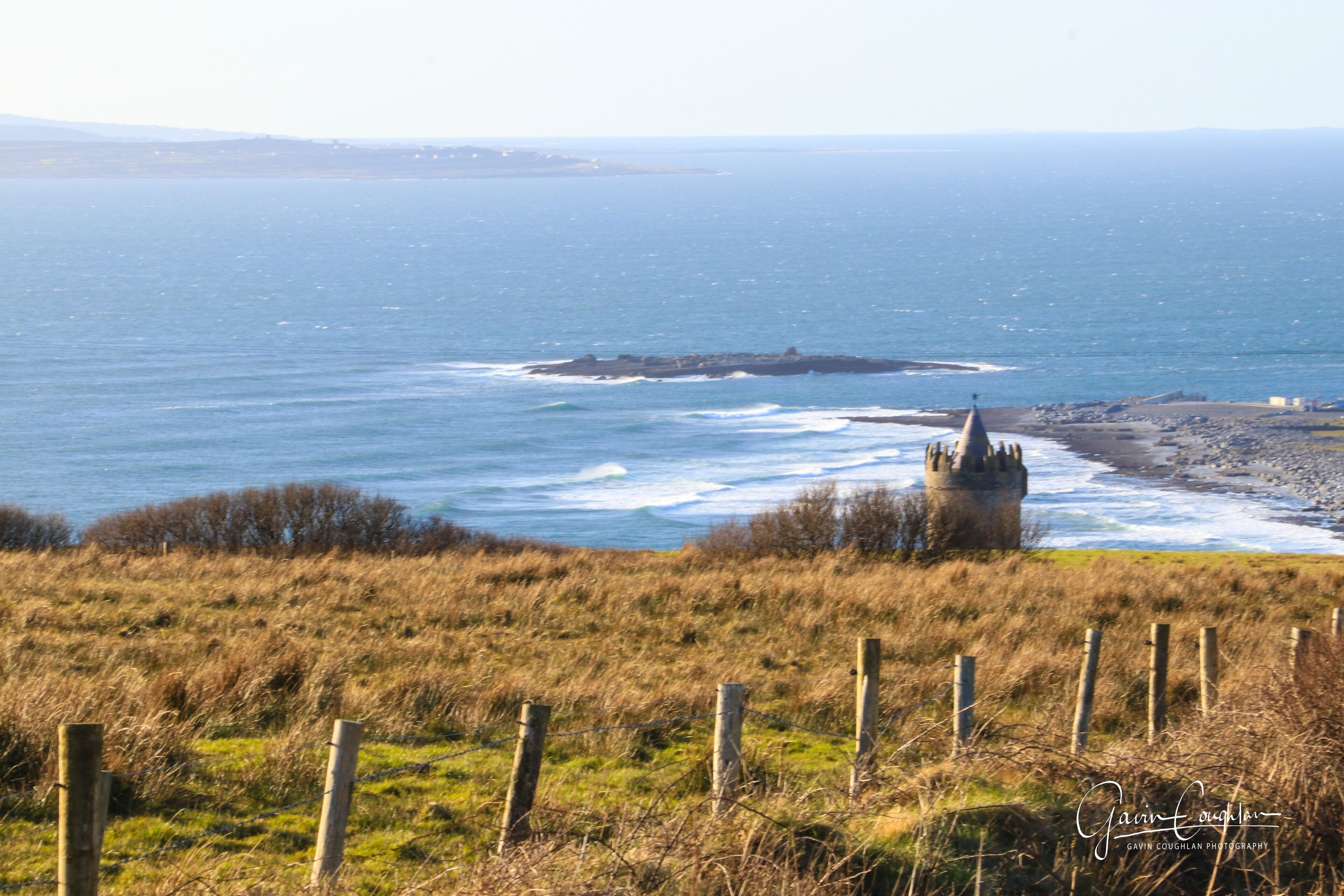 A coastal landscape with a grassy foreground, a wooden fence, and a distant view of the sea and a small rocky structure.