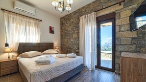 Une chambre avec un mur en pierre, un lit recouvert d’une courtepointe, une table de chevet, une lampe et une vue sur l’extérieur, visible à travers une porte vitrée.
