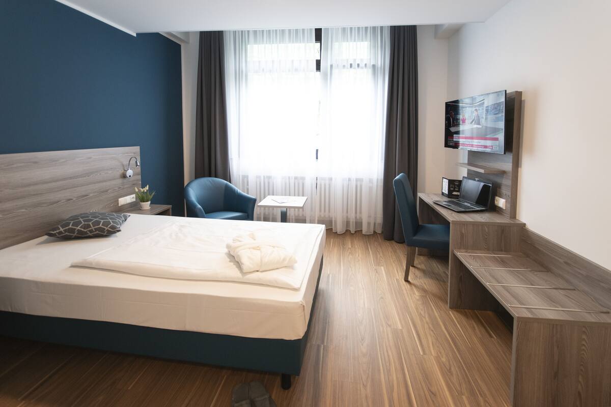 A hotel room with a bed, a desk, a chair, a TV, and a window with curtains.
