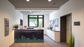 A reception desk with a cityscape graphic, a window with a view of trees, and a door to the right. - New Work Hotel Essen (Essen)