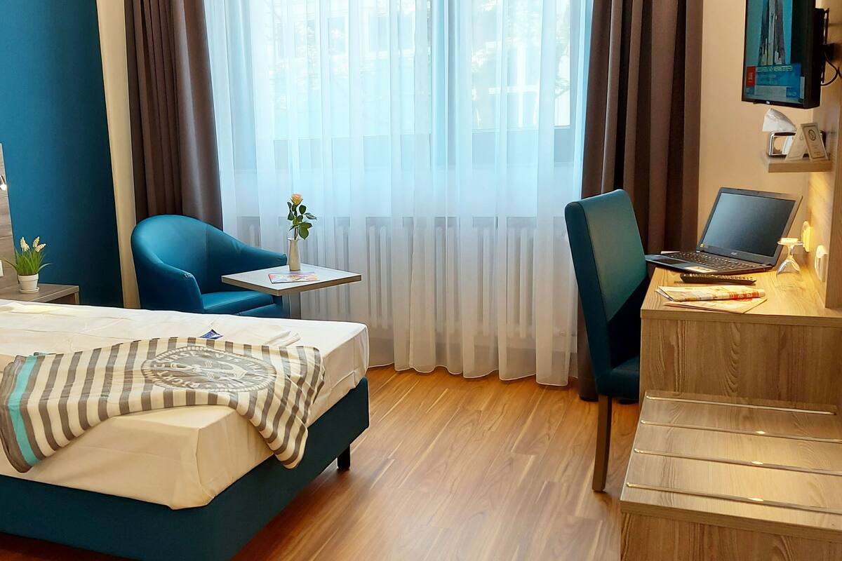 A hotel room with a bed, a desk with a laptop, a chair, a small table with a plant, and a window with curtains.
