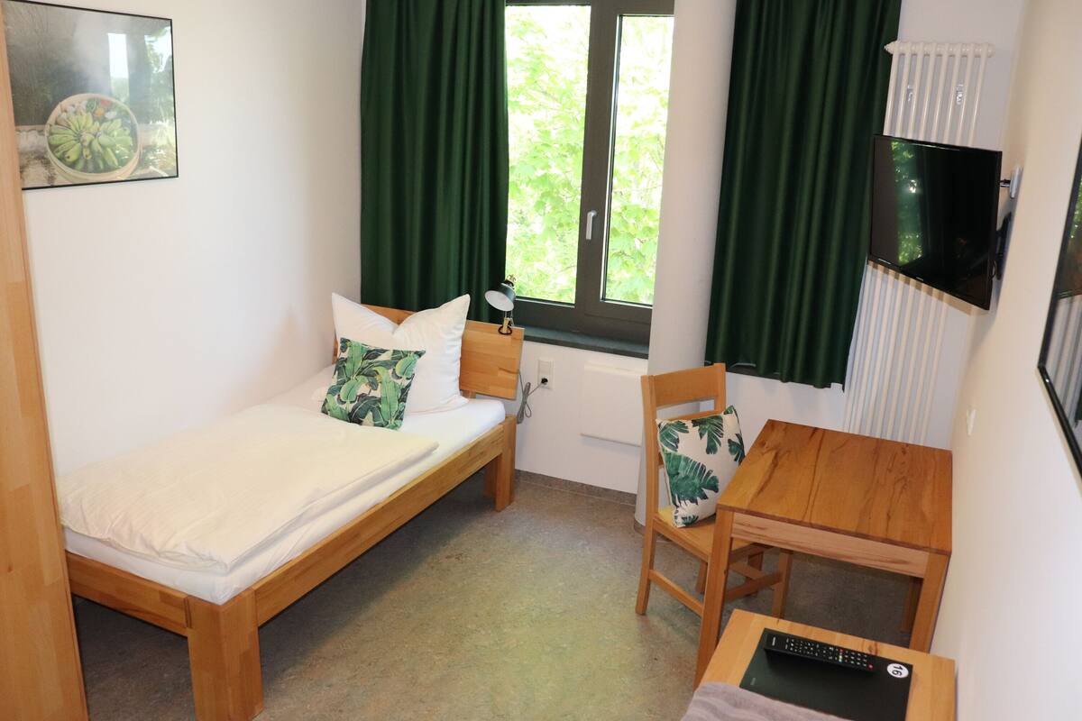 A small, tidy room with a single bed, a wooden table, a chair, a TV, and a window with curtains.