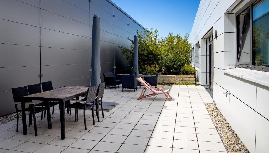 Un patio al aire libre con una mesa de comedor y sillas, una silla de descanso, y un edificio moderno con grandes ventanales.
