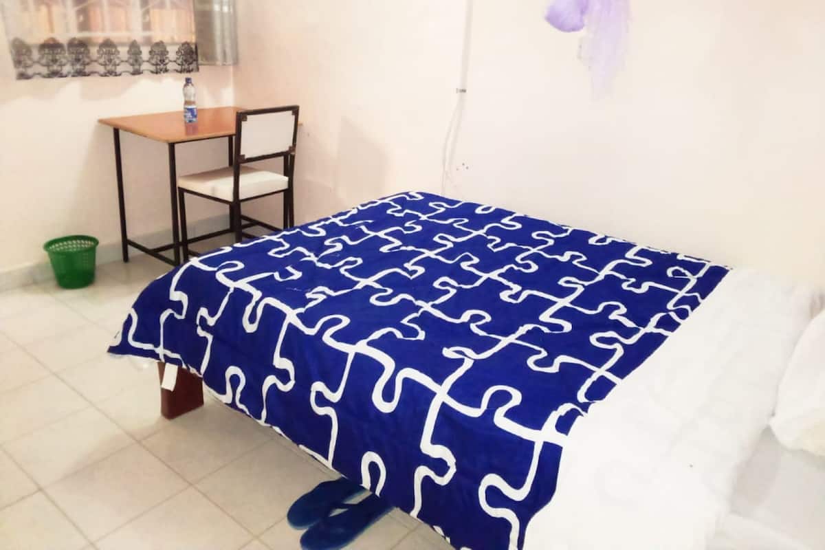 A room with a bed, a chair, a table, a water bottle, and a green basket.