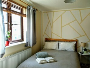 Un dormitorio con cama, ventana con persianas, radiador, mesita de noche con lámpara y pared con diseños geométricos.