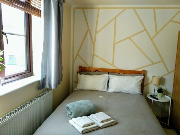 Una habitación de hotel con cama, radiador, ventana con cortinas, mesita de noche con lámpara y pared con diseños geométricos.