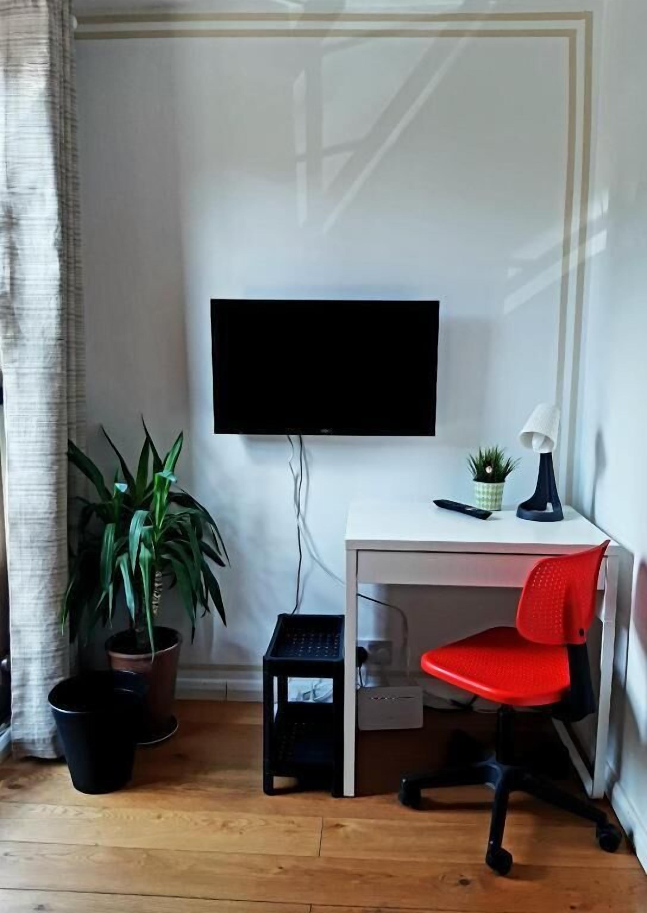 Un espacio de oficina pequeño y ordenado con un escritorio blanco, una silla de oficina roja, un monitor negro, una pequeña planta en maceta y un teclado negro sobre un piso de madera.