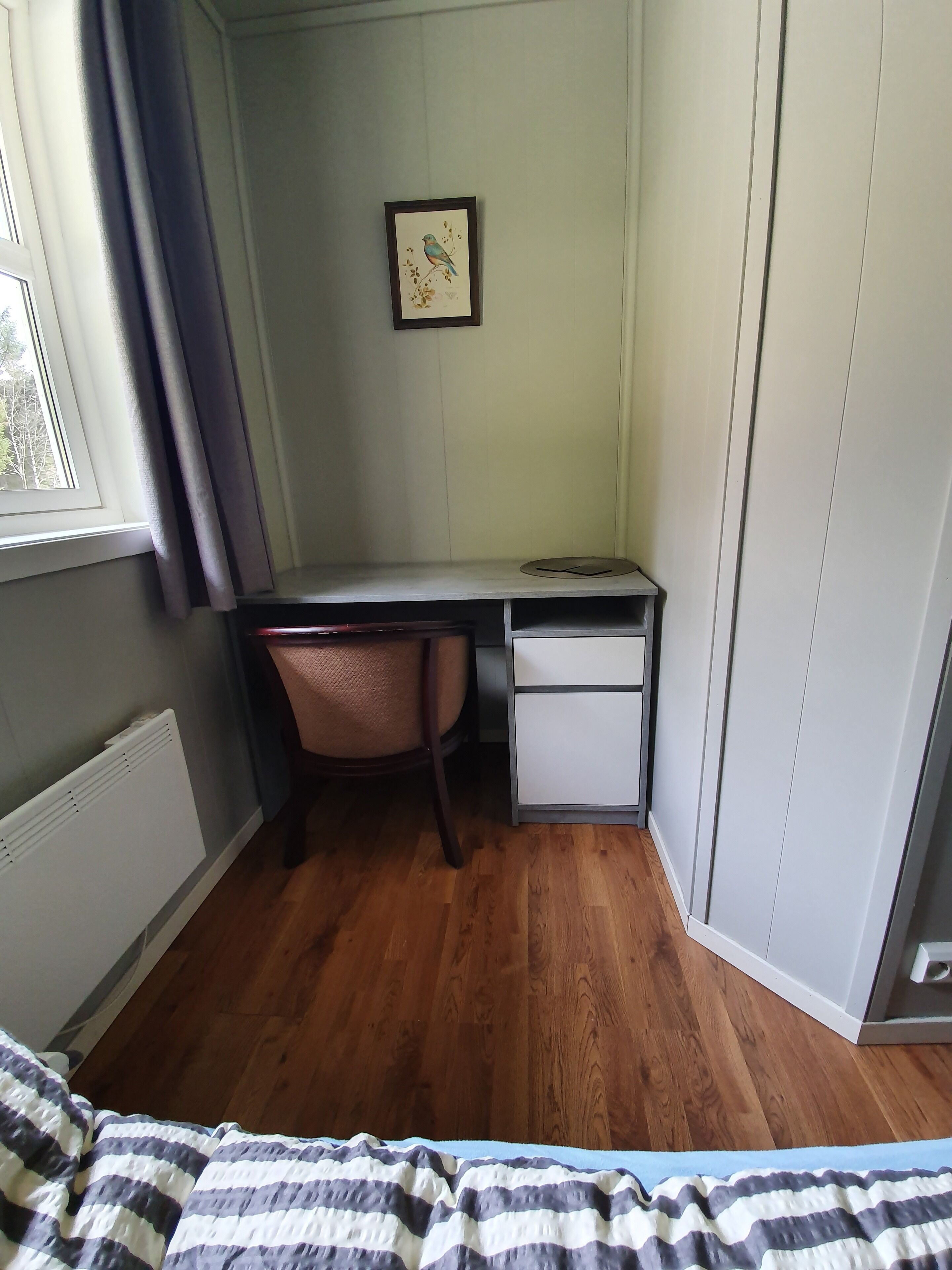 Um quarto com cama, escrivaninha, cadeira, radiador e janela com cortinas.