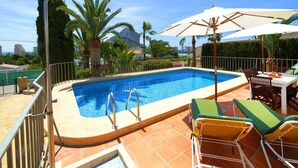 A swimming pool with a clear blue water, surrounded by a tiled deck, and equipped with a metal ladder. There are lounge chairs and a table under a large umbrella. - Stunning private villa for 6 guests with A/C, private pool, WIFI, TV, terrace and pets allowed (Calpe)