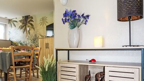 Uma sala de jantar com mesa e cadeiras de madeira, um vaso com flores azuis, um besouro decorativo e uma estante com figurines de elefante.